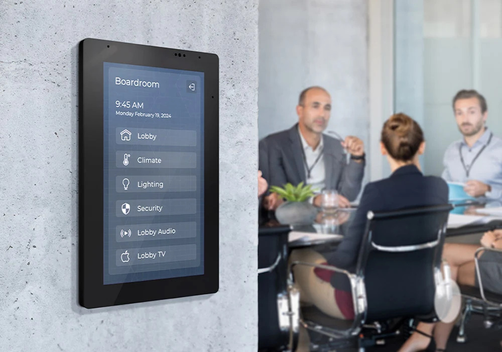 Wall-mounted touch control panel in a boardroom with people meeting in the background