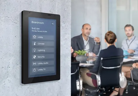 Wall-mounted touch control panel in a boardroom with people meeting in the background